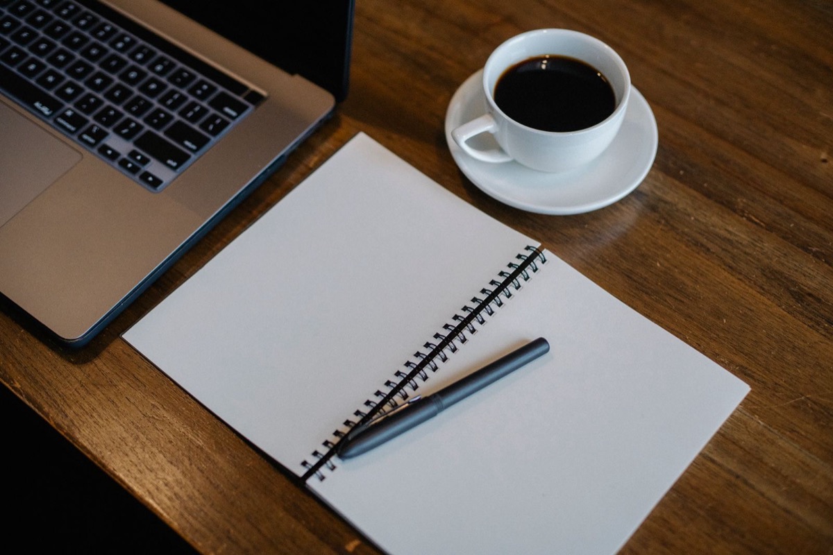 A laptop, an open blank spiral notebook with a pen and a cup of black coffee on a wooden desk