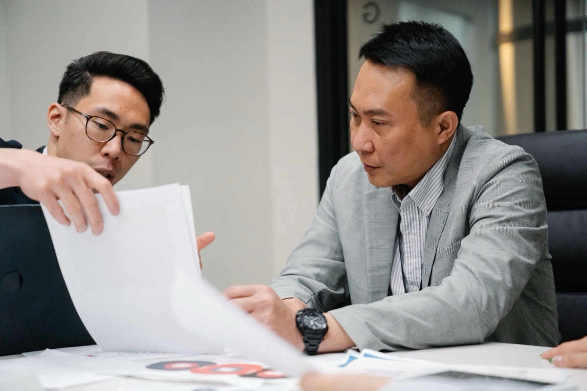 Two men leaning over printed pages together in a meeting room, with a laptop and charts on the table