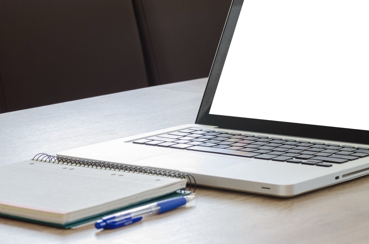 A laptop with a blank screen beside a spiral notebook and a blue pen on a wooden desk