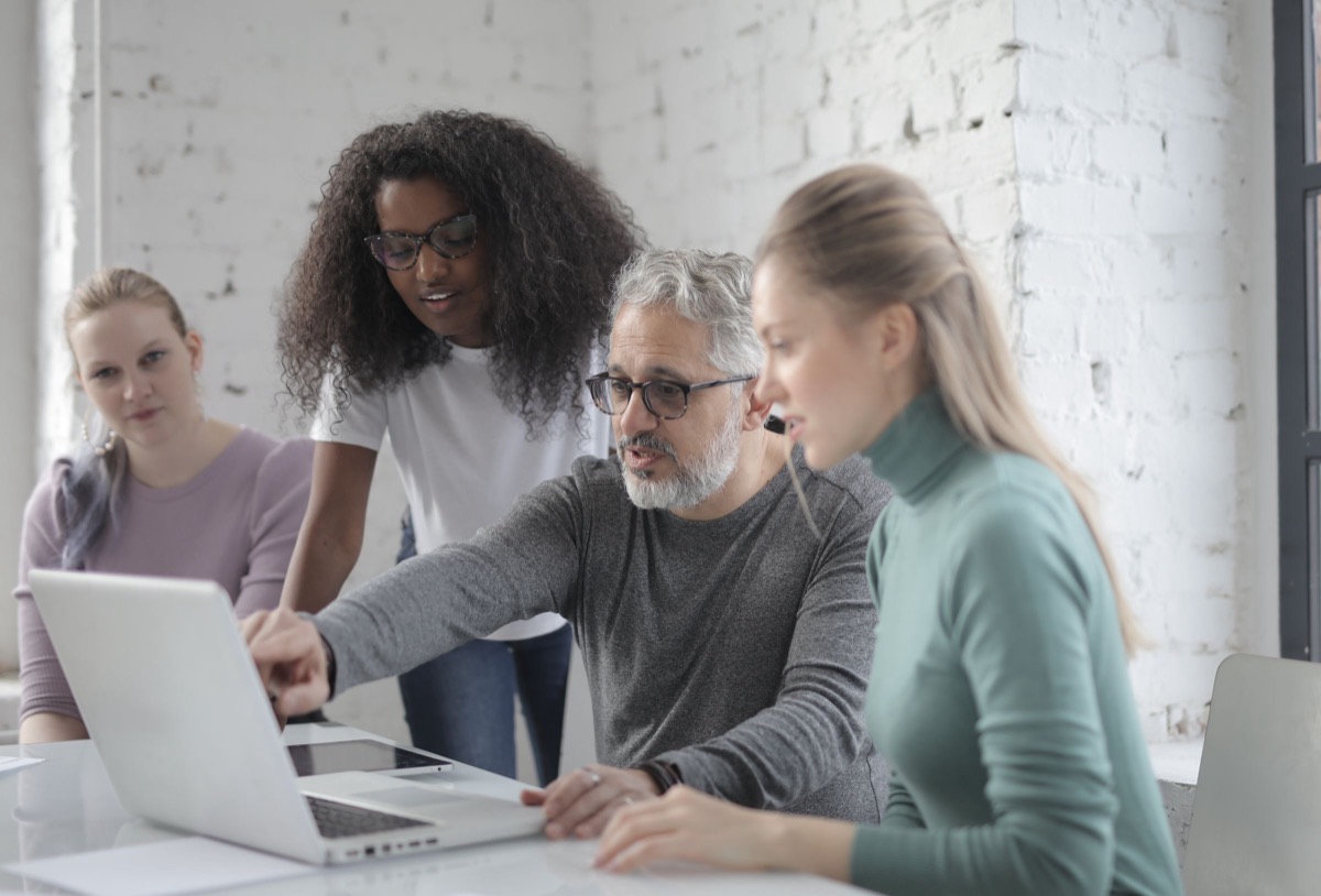 Four colleagues gathered around a laptop at a table in a bright office with a white brick wall