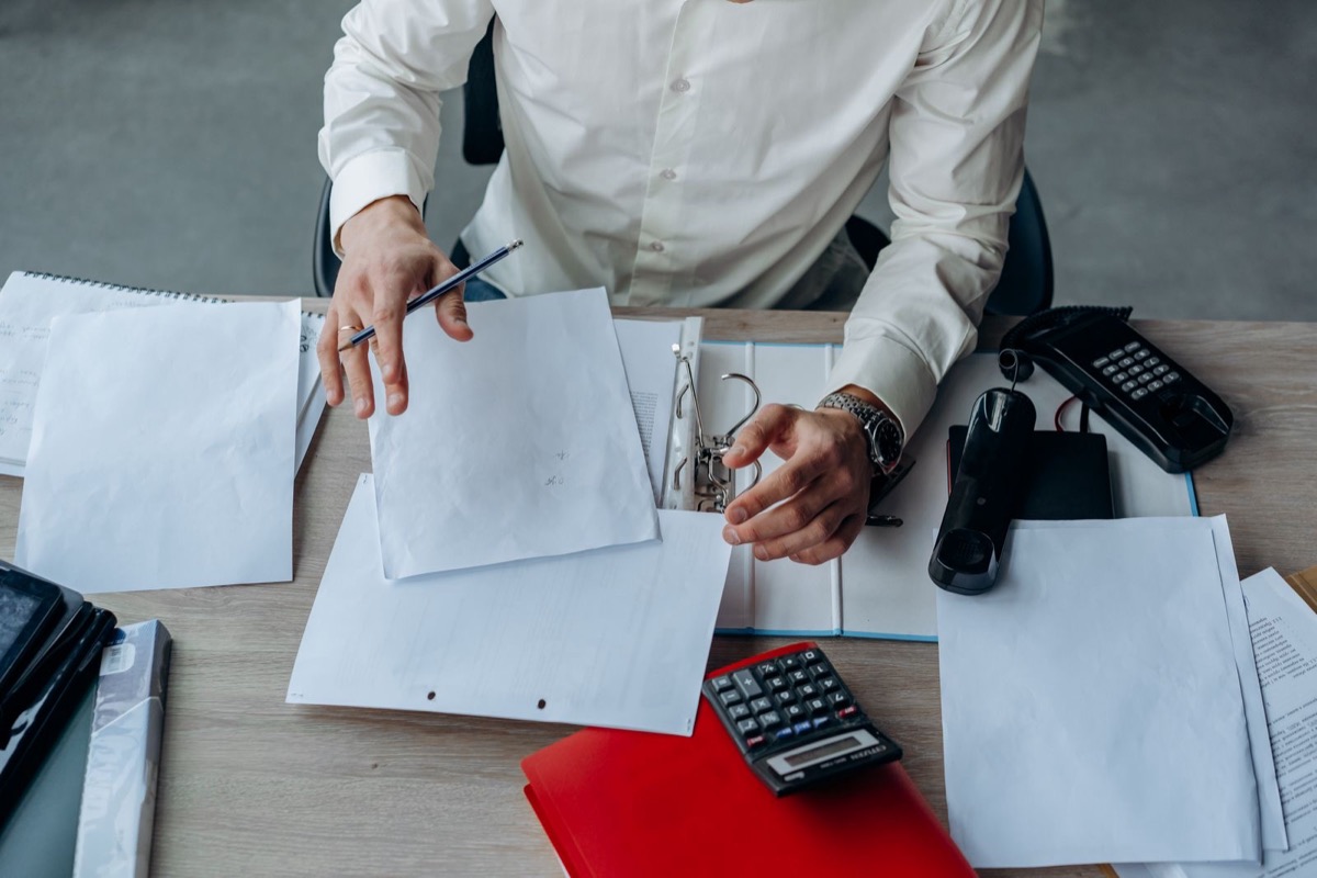 A person at an office desk sorting loose sheets of paper and a ring binder beside a calculator and telephone
