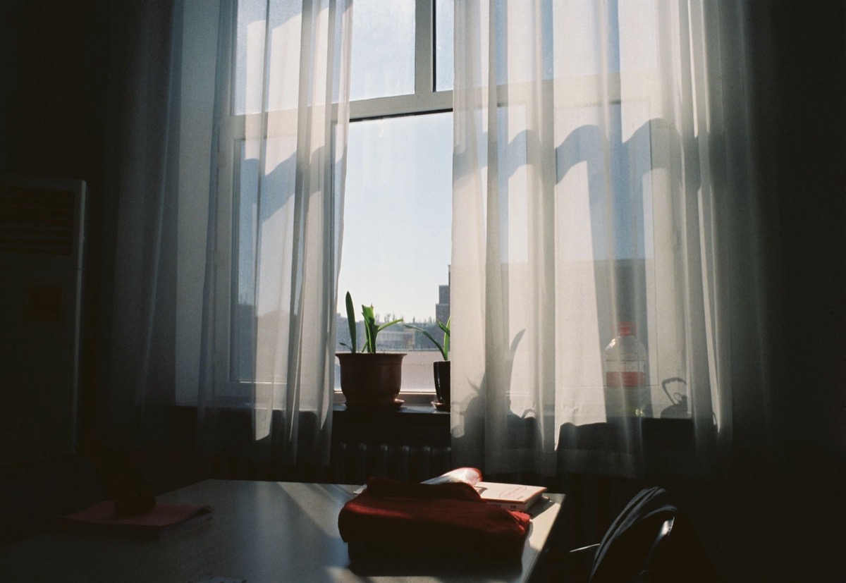 Sheer curtains at a tall window in a dim room, with potted plants on the sill and a desk
