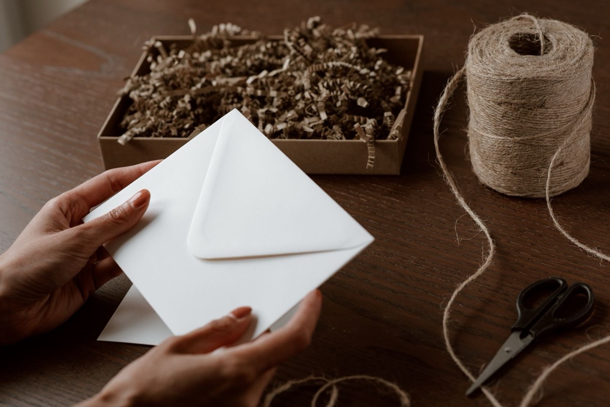 Hands holding a plain white envelope above a wooden table with a box of paper shred, twine and scissors