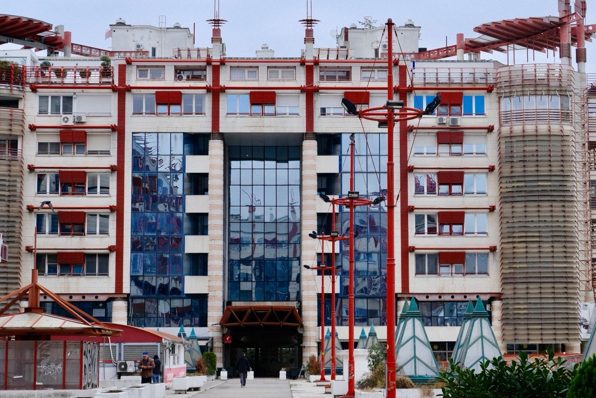 A modern multi-storey building with a glass central entrance and red steel posts, seen across a paved forecourt
