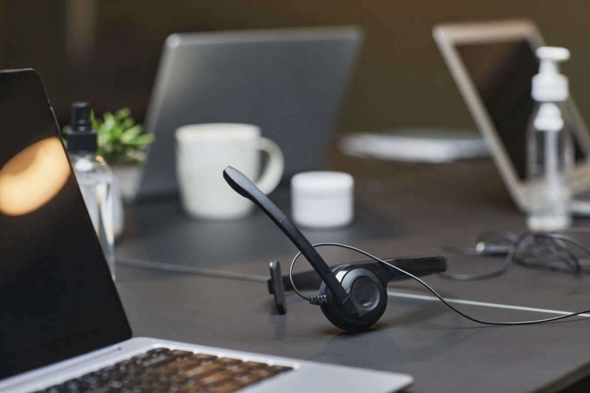 A headset lying on a dark desk among open laptops, a white mug and bottles of hand sanitiser