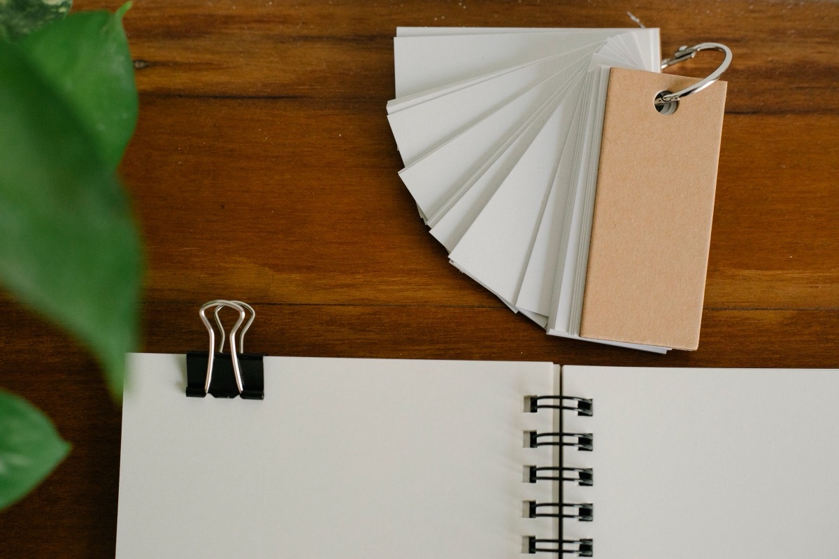 An open blank spiral notebook with a black binder clip and a fan of blank cards on a wooden desk