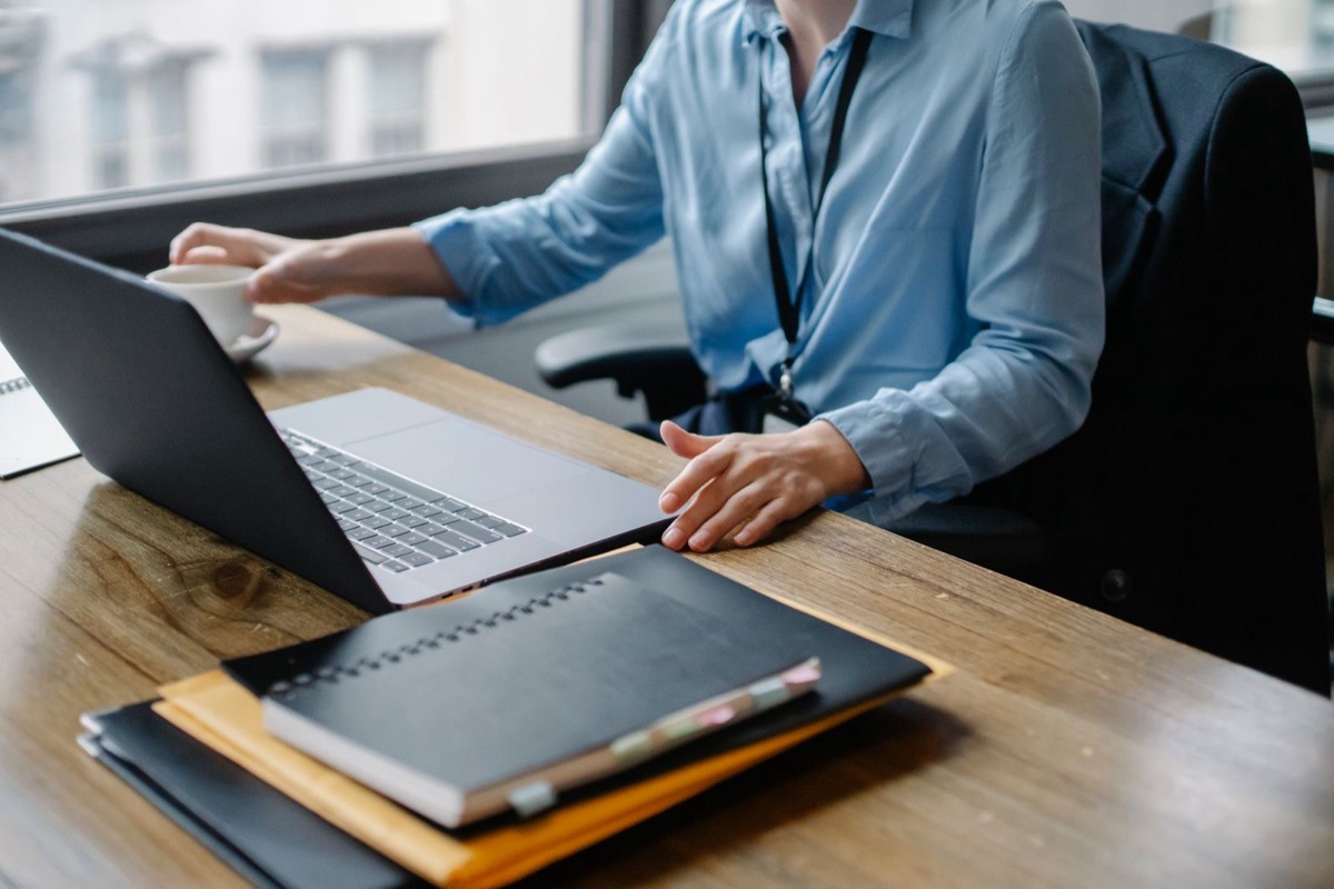 A person in a blue shirt at a laptop on a wooden desk, with folders and a notebook nearby
