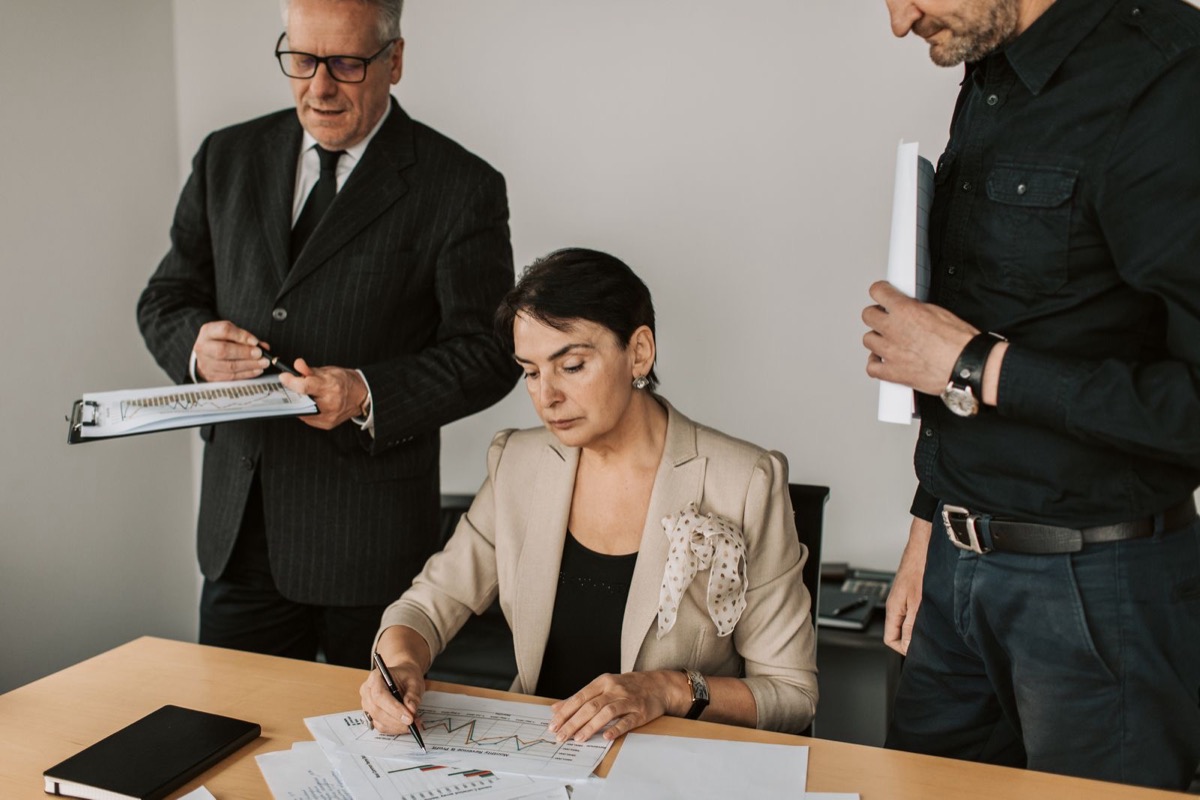 A seated woman marking printed charts at a desk while two colleagues stand beside her with papers