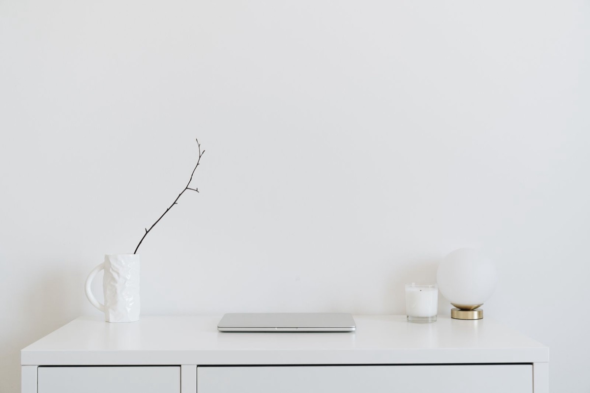 A closed silver laptop on a white chest of drawers beside a jug with a twig and a round lamp