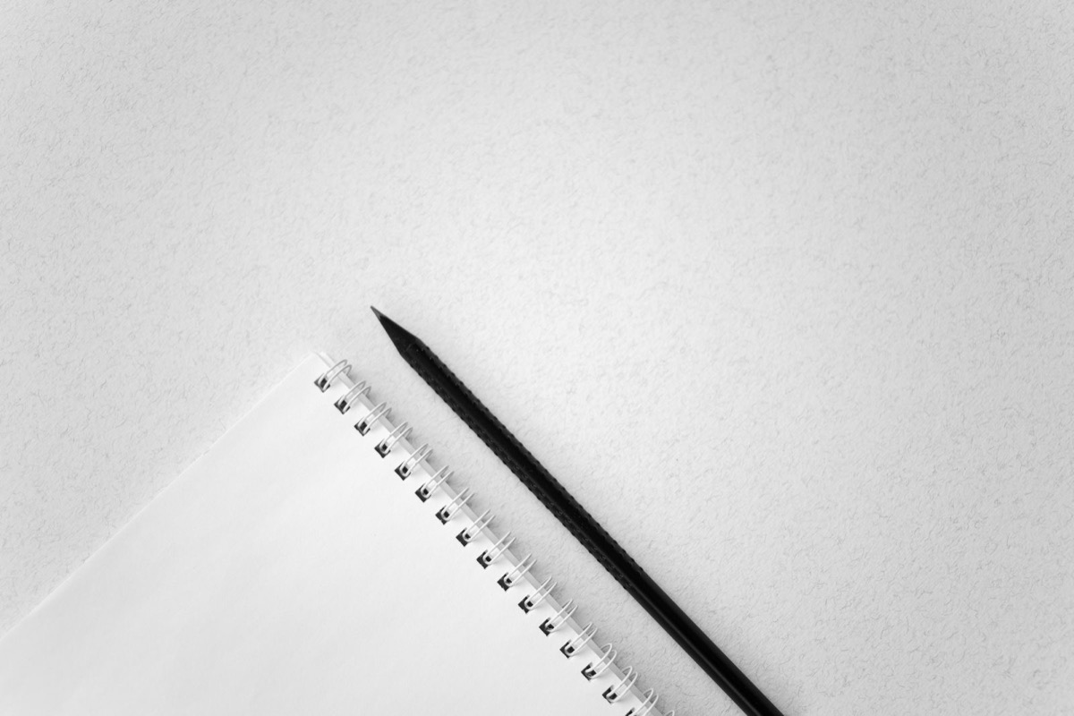 A black pencil beside a blank spiral-bound notebook on a pale grey desk surface