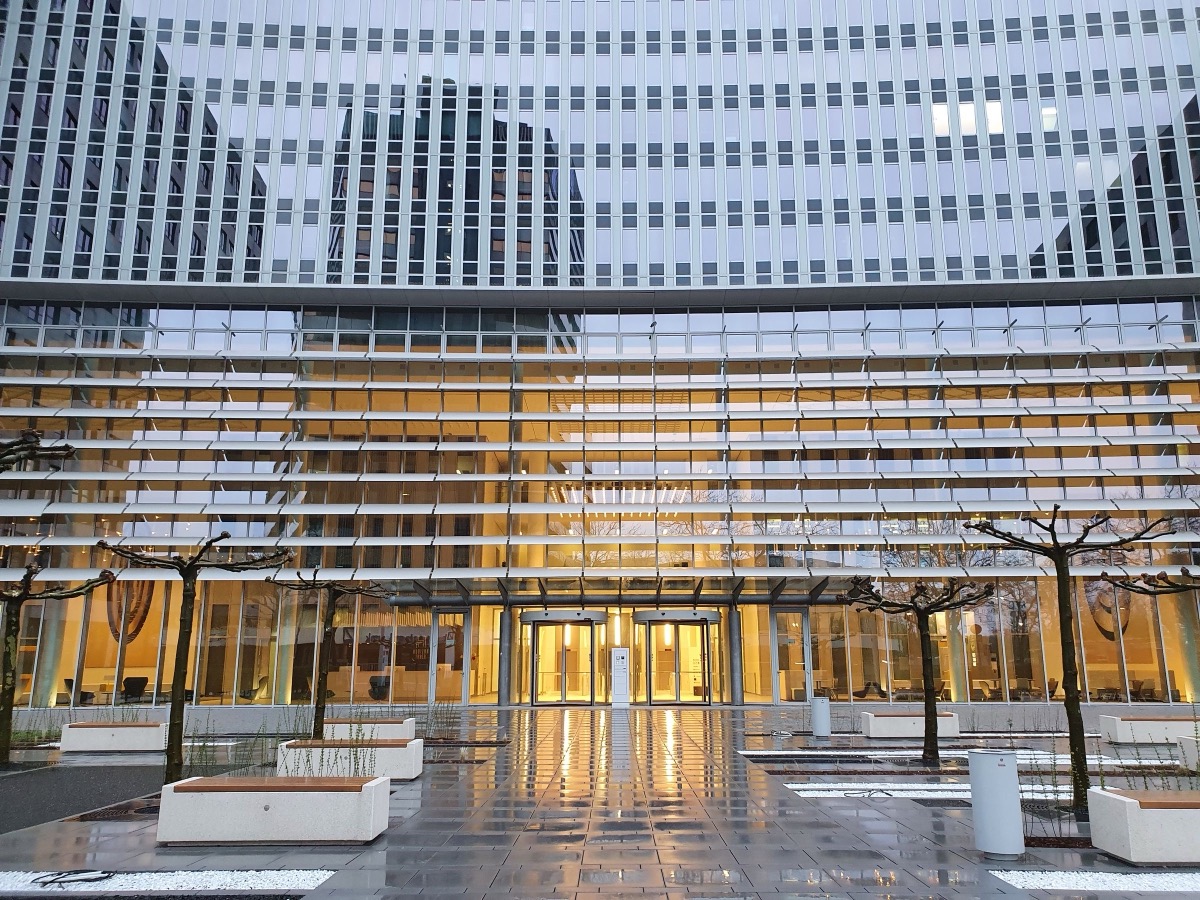 A glass-fronted office building with a lit entrance, a paved forecourt, planters and bare trees